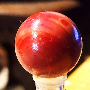 May include: A spherical object with a glossy, smooth surface featuring a deep red color with subtle, swirling lighter red and orange patterns. The sphere is mounted on a clear, cylindrical base that tapers slightly toward the top.