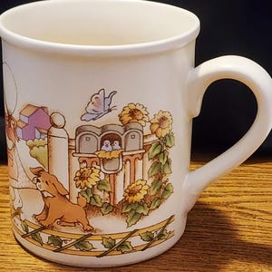 Może przedstawiać: Biały kubek ceramiczny z kolorową ilustracją domu, psa, motyla i słoneczników. Kubek ma uchwyt po prawej stronie.