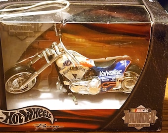 Pode incluir: Uma motocicleta Hot Wheels NASCAR Thunder Rides de fundição de metal prateada e azul com a marca Valvoline. A motocicleta está em uma vitrine de plástico transparente.