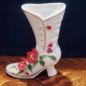 May include: White ceramic boot-shaped vase with red and green floral details. The vase has a gold trim and is decorated with red flowers and green leaves.
