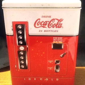 May include: A vintage Coca-Cola metal container designed to resemble an ice-cold bottle vending machine. The container is red and white with the Coca-Cola logo and the words "Drink Coca-Cola in Bottles". The words "Ice Cold" are at the bottom.