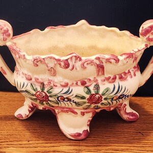 Puede incluir: Un macetero de cerámica blanca con diseños florales rosas y verdes. El macetero tiene dos asas ornamentadas y tres patas.