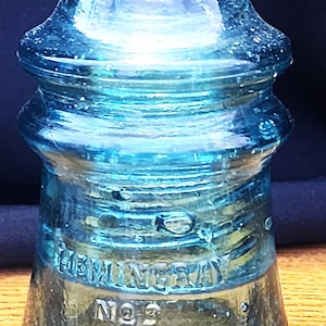May include: A vintage blue glass electrical insulator with a rounded top and a flared base. The glass features embossed text that reads HEMINGRAY and NO 9. The surface of the translucent blue glass shows small air bubbles and a smooth, glossy finish.