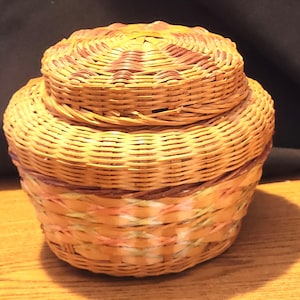 May include: A round, lidded basket made of woven light brown and reddish-brown material. The lid has a star-like design. The basket's base features a band of pink, yellow, and brown woven strands. The basket is sitting on a wooden surface.