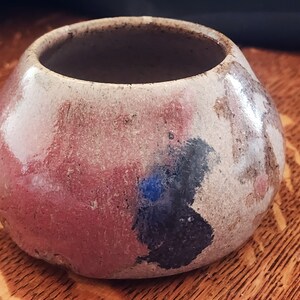 Op de afbeelding: Een kleine, ronde keramische pot met een brede opening. De pot is geglazuurd in tinten roze, grijs en zwart, met een blauwe accent.