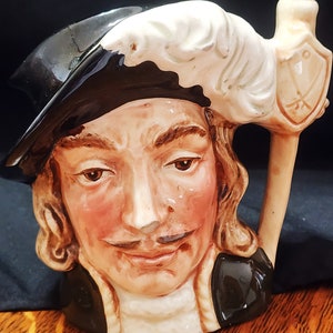 Könnte beinhalten: Eine Keramiktasse in Form eines Männerkopfes, der einen schwarzen Hut mit einer weißen Feder trägt. Die Tasse hat ein braunes und weißes Design und einen Griff in Form eines Stabes.