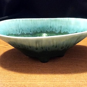 Puede incluir: Un cuenco grande de cerámica de forma ovalada con un esmalte verde y azul. El cuenco tiene una abertura ancha y poco profunda y un fondo redondeado. El esmalte es desigual, creando un efecto moteado.