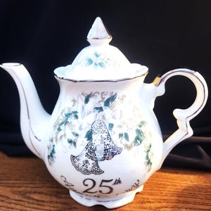 May include: White porcelain teapot with a lid, spout, and handle. The teapot is decorated with teal floral patterns and a bell design. The number "25th" is printed on the front. Gold trim accents the edges.