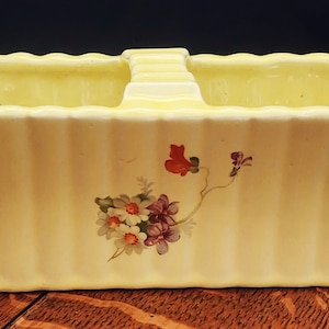 May include: A yellow ceramic planter with a floral design. The planter has a handle and is divided into two sections.