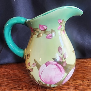 Op de afbeelding: Een groene en witte keramische kan met een bloemenmotief. De kan heeft een groene handgreep en een parelmoerglans. De bloemen zijn roze en wit.