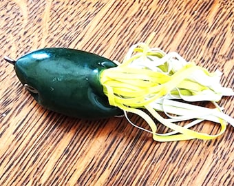 Può includere: Un'esca da pesca verde scuro con un corpo liscio e arrotondato e un amo in metallo sulla parte anteriore. L'estremità posteriore dell'esca presenta un gruppo di lunghi e sottili fili di gomma giallo brillante che si estendono verso l'esterno, creando un aspetto a frange.