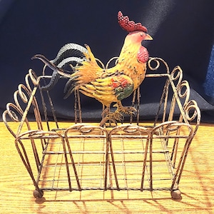May include: A decorative metal rooster figurine with a red comb and yellow body stands in a wire basket. The basket has a distressed gold finish and is supported by small ball feet. The rooster has colorful accents.