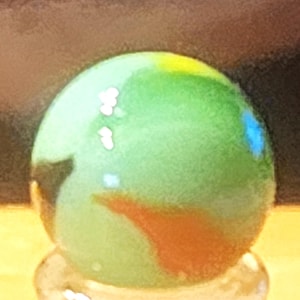 May include: A spherical glass marble featuring a translucent light green base with swirling patterns of opaque white, bright yellow, and a distinct patch of burnt orange. The surface is smooth and reflective, catching light to create bright highlights.
