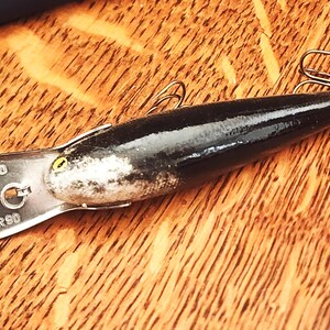 May include: A black and silver Rapala Deep Diver 90 fishing lure with a yellow eye and a metallic diving lip. The lip is clearly engraved with the text RAPALA, FINLAND, and DEEP DIVER 90. Two metal treble hooks are attached to the body of the lure.