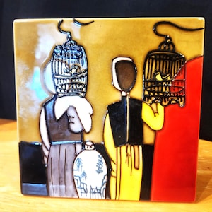 May include: Square ceramic tile featuring a stylized illustration of two figures holding birdcages. The figures are depicted in black, yellow, and brown, with a gold and red background. A white and blue vase is at the bottom. The tile is approximately 6 inches square.