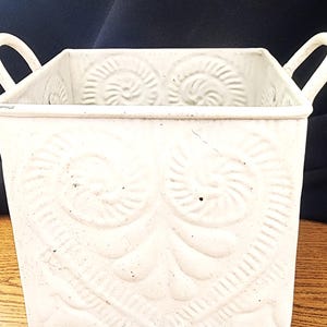 Puede incluir: Una maceta de metal blanca y cuadrada con dos asas. La maceta presenta un diseño en relieve de patrones arremolinados. La maceta está pintada de blanco y se encuentra sobre una superficie de madera.
