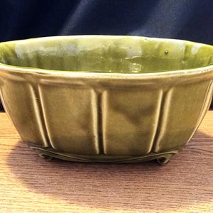 Puede incluir: Una maceta de cerámica verde con un diseño acanalado y tres pequeños pies. La maceta tiene forma ovalada y un acabado brillante.