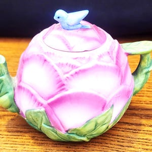 May include: A decorative ceramic teapot shaped like a pink rose, with green leaves and a light blue bird on the lid. The teapot has a curved handle and spout, and is set on a wooden surface.