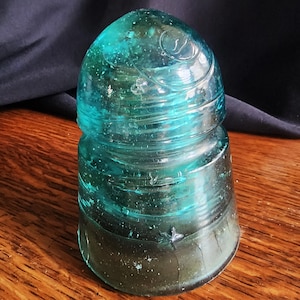 May include: A vintage teal-colored glass insulator with a rounded top and a cylindrical base. The glass has a textured surface and a swirl design on the top. The insulator is sitting on a wooden surface.