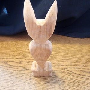 Puede incluir: Una talla de madera de un gato estilizado con orejas grandes. La talla está hecha de madera de color claro y tiene un acabado liso.