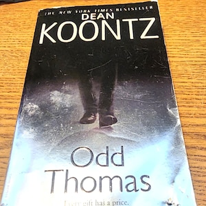 May include: A paperback copy of Dean Koontz's "Odd Thomas". The cover is predominantly dark, with the author's name and title in white. The tagline "Every gift has a price" is printed at the bottom.