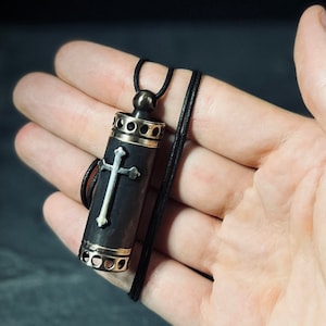 Puede incluir: Un collar con un colgante de cruz negro y plateado, con un encanto cilíndrico. El encanto es de madera y tiene una cruz plateada en la parte delantera.