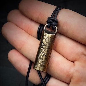 May include: A brass pendant necklace with a textured cylindrical charm hanging from a black leather cord.