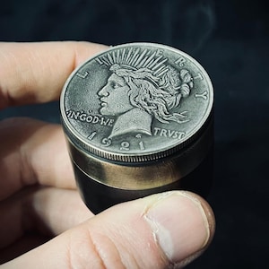 May include: A cylindrical metal grinder featuring a silver-toned 1921 Peace dollar coin on the top lid. The coin displays the profile of Lady Liberty with a radiant crown, the text LIBERTY, IN GOD WE TRVST, and the year 1921. The base is dark metal.