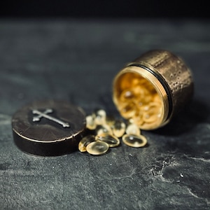 Peut inclure: Un petit récipient rond en métal avec une croix sur le couvercle est ouvert, laissant échapper de petites capsules rondes et transparentes. Le récipient et les capsules sont sur une surface gris foncé.