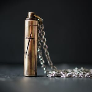May include: A silver chain necklace with a brass pendant shaped like a cylinder. The pendant has a cross carved into it.