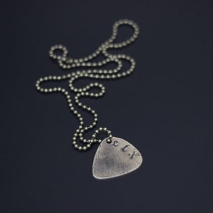 Könnte beinhalten: Eine silberne Halskette mit einem silbernen Gitarren-Plektrum-Anhänger. Das Plektrum trägt die eingravierten Buchstaben "ETX".