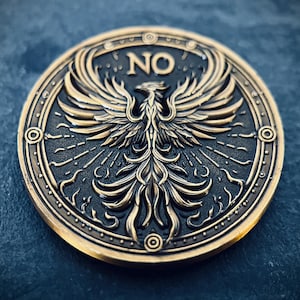 Może przedstawiać: Złota moneta metalowa z detalicznym wzorem feniksa z rozpostartymi skrzydłami. Moneta ma czarne tło i napis "NO" wygrawerowany w środku.