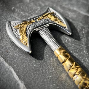 May include: Close-up of a silver and gold axe pendant. The axe head features a detailed gold eagle design on a silver background. The handle is silver with a gold-colored, textured grip. The axe is a miniature decorative item.