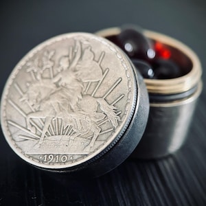 May include: A silver container with a lid featuring a detailed relief of a figure with outstretched arms, with the year 1910. The container is open, revealing dark, round objects inside. The container is on a dark surface.
