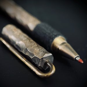 Könnte beinhalten: Nahaufnahme eines silbernen Stifts mit schwarzem Gummibeschlag und roter Spitze. Der Stift liegt neben einer silbernen Metall-Stiftkappe mit gehämmerter Textur.