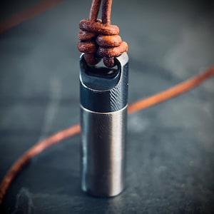May include: A silver-colored, cylindrical pendant with a textured surface, suspended from a brown leather cord. The cord is knotted at the top, and the pendant has a curved opening. The background is a dark, blurred surface.