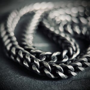 May include: Close-up of a silver curb chain necklace. The chain features thick, interlocking links, creating a bold, textured appearance. The image focuses on the chain, with a blurred background, highlighting the jewellery's details.