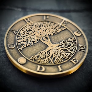 Puede incluir: Una moneda de metal dorado con un diseño de árbol de la vida. El árbol está rodeado por las palabras "Carpe Diem" en un círculo.