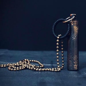 Puede incluir: Un colgante de metal oscuro con una superficie texturizada cuelga de una cadena dorada. La cadena está hecha de pequeñas cuentas redondas. El colgante está unido a la cadena con un cordón negro.
