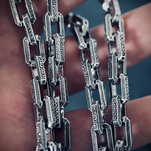 May include: A silver chain necklace with intricate, detailed designs on each link. The chain is made of multiple strands, creating a layered effect.