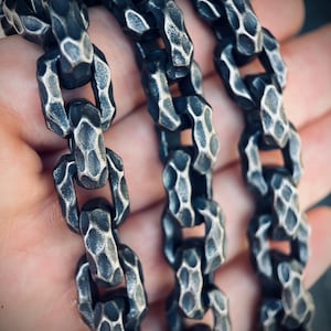 Op de afbeelding: Een zware metalen ketting met dikke, in elkaar grijpende schakels met een donkere, geoxideerde afwerking en een opvallend gehamerd, getextureerd oppervlak. De individuele schakels zijn rechthoekig van vorm en hebben een robuuste uitstraling.