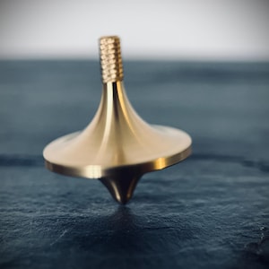 May include: A gold-colored spinning top with a textured top and a rounded, conical body. The top is set against a dark, textured surface, creating a contrast that highlights its shape and metallic sheen. A classic toy.