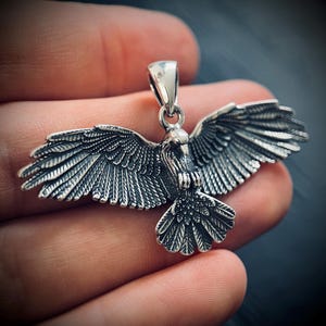 Pode incluir: Um pingente de prata detalhado em forma de pássaro com as asas abertas. O pingente tem detalhes intrincados de penas e uma argola na parte superior para uma corrente. O pássaro é retratado em uma pose dinâmica, sugerindo voo.