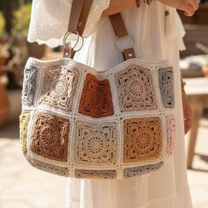 May include: A large, handmade crochet tote bag with a patchwork design. The bag features squares in various colors, including beige, brown, orange, and gray. It has brown leather handles attached with silver rings, and is carried by a person wearing a white dress.