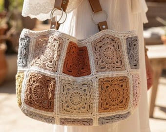 May include: A large, handmade crochet tote bag with a patchwork design. The bag features a variety of neutral colors, including beige, brown, and gray, arranged in square patterns. It has brown leather handles attached with silver rings, and is carried by a person wearing a white dress.