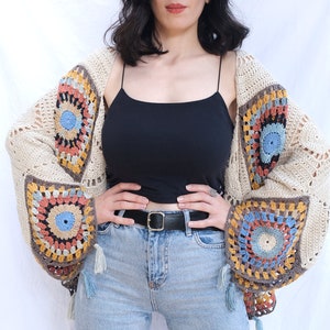 May include: A crocheted cardigan with a cream background and colorful geometric patterns. The cardigan has a loose fit and long sleeves with tassels.