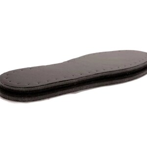 May include: A black leather insole with a layer of foam padding. The insole has a stitched edge and a textured surface.