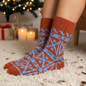 Puede incluir: Un par de calcetines de punto en color óxido con un patrón geométrico azul claro. Los calcetines están sobre una alfombra blanca, con un árbol de Navidad y velas al fondo.