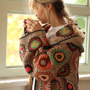 Könnte beinhalten: Eine braune Häkeljacke mit Kapuze, die mit bunten kreisförmigen Mustern in Rot-, Grün-, Gelb- und Blautönen verziert ist.