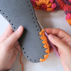 May include: A person is crocheting a bright orange crocheted edge onto a gray leather sole. The crocheted edge is being attached to the sole with a pink crochet hook.